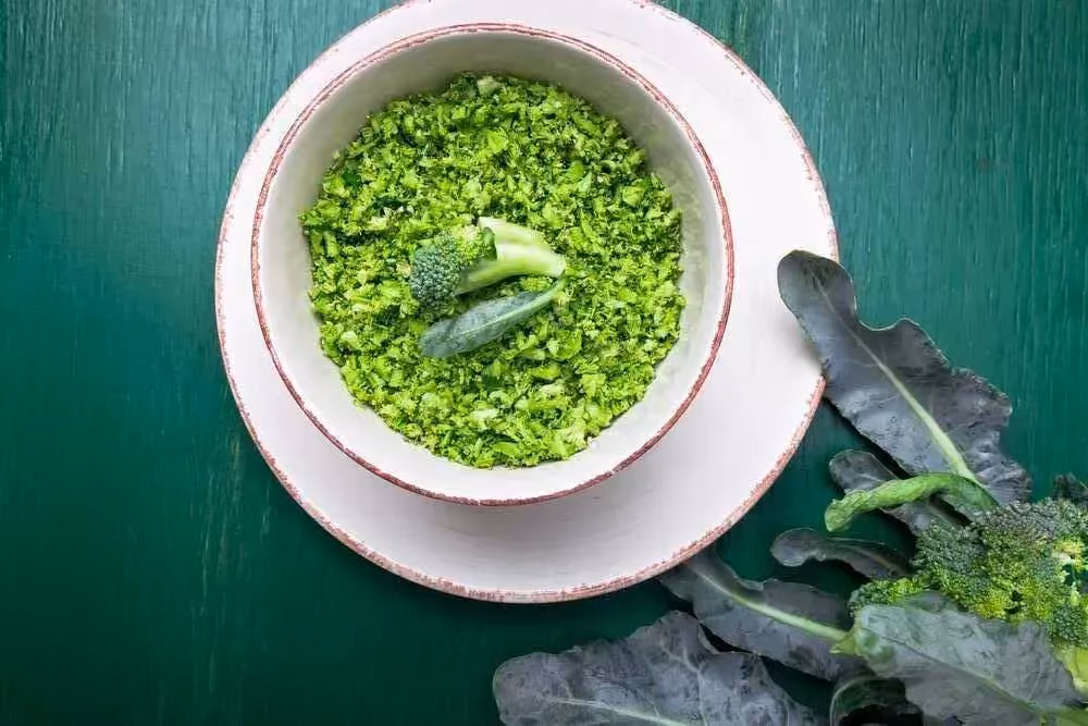 In a white earthenware bowl, there is broccoli chopped to the size of rice grains. Two small broccoli florets are placed on top.