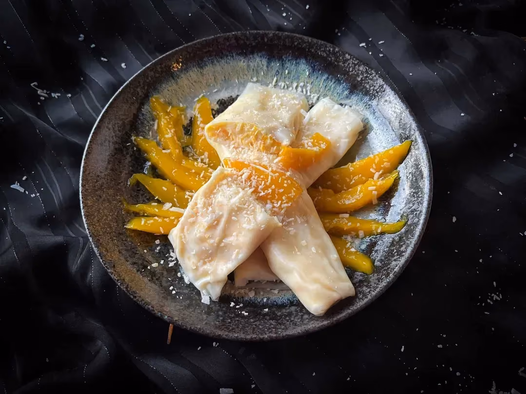 In a small bowl, there are four spring rolls served with thin slices of mango, topped with coconut.