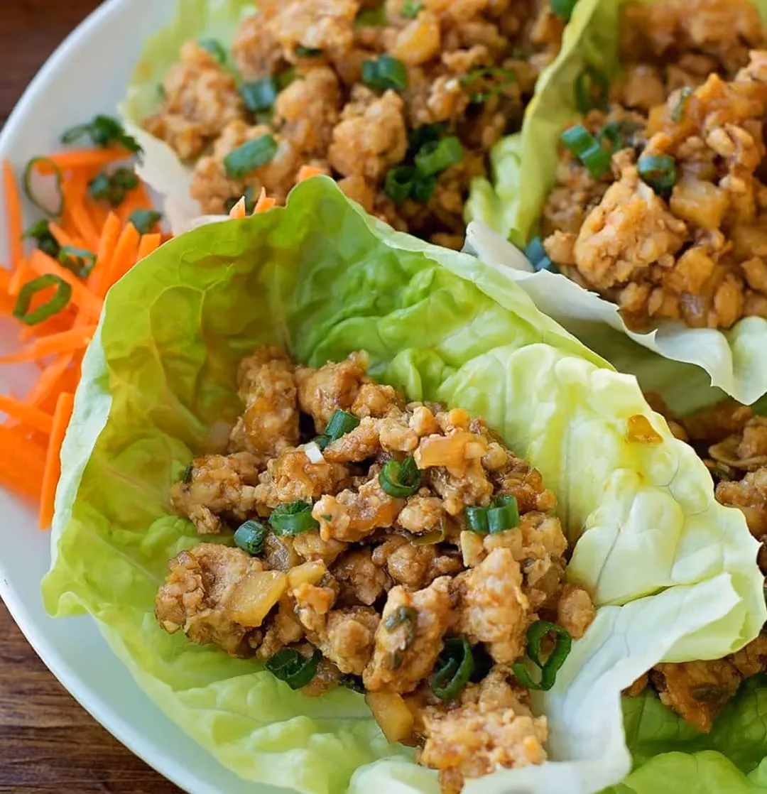 Dans un plat blanc, se trouve des feuilles de laitue contenant des petits morceaux de poulet façon wraps.