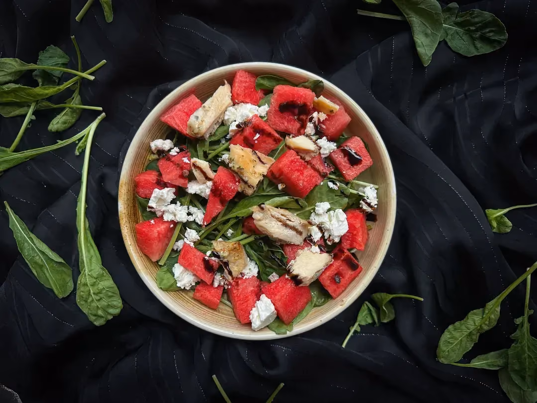 The watermelon, arugula, and feta salad is presented on a white plate. Five small slices of watermelon are placed on the side.