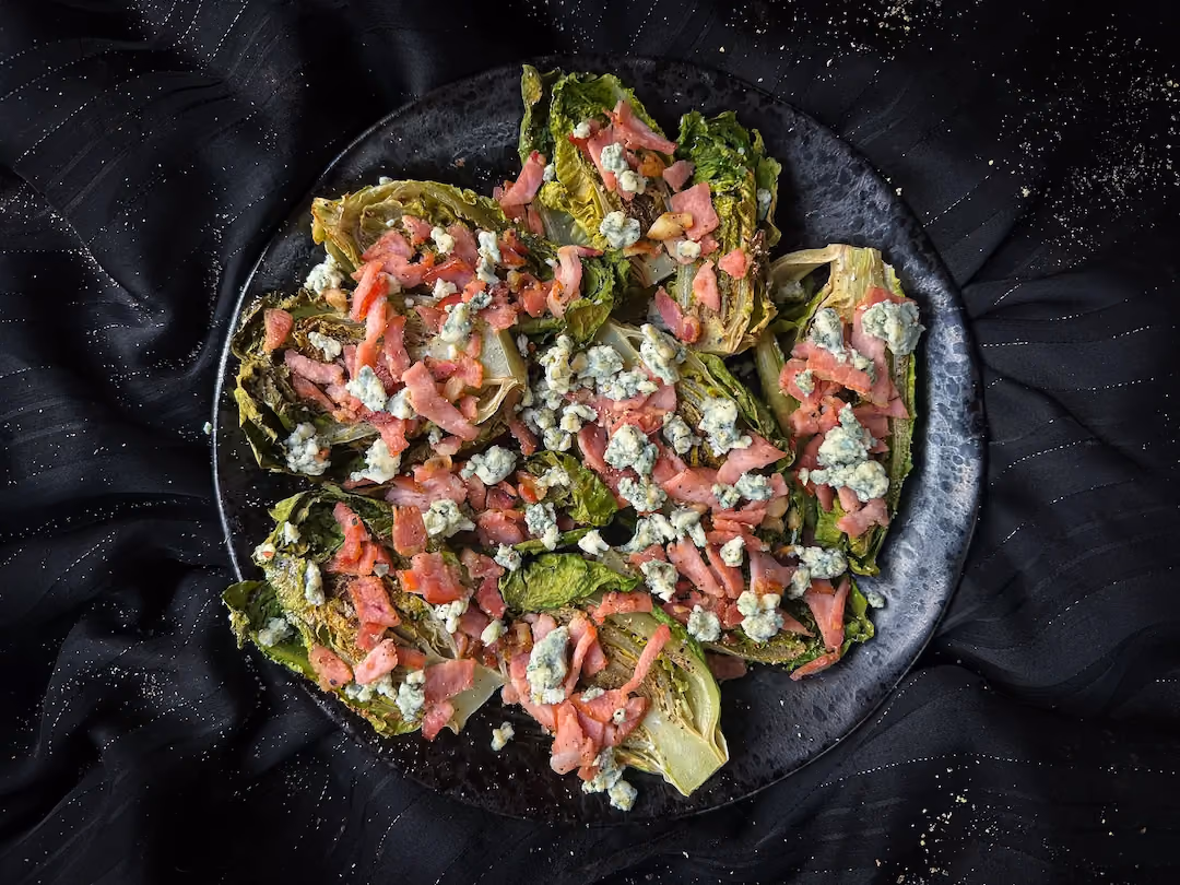 On a black plate is grilled romaine salad with bacon and gorgonzola.