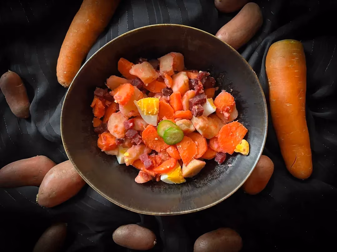 Un petit saladier accueille la salade russe. Autour du plat se trouve deux carottes et des pommes de terre rosées.