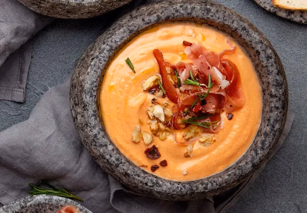 In a bowl, the slightly yellow-orange Salmorejo is sprinkled with small pieces of ham and egg.