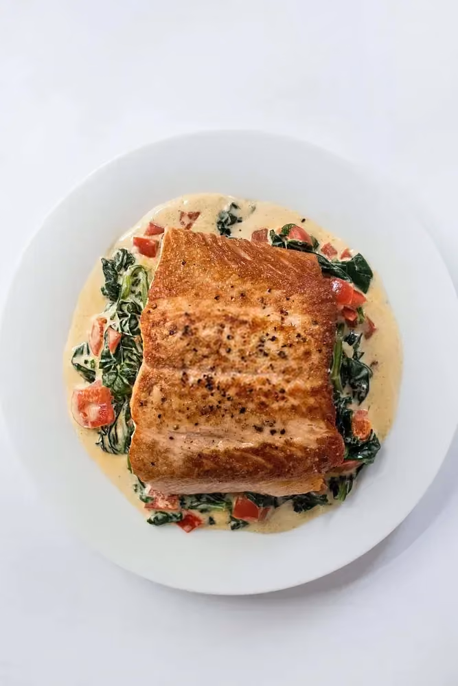 On a white plate, salmon fillets with the skin on are served over a Tuscan lemon sauce.