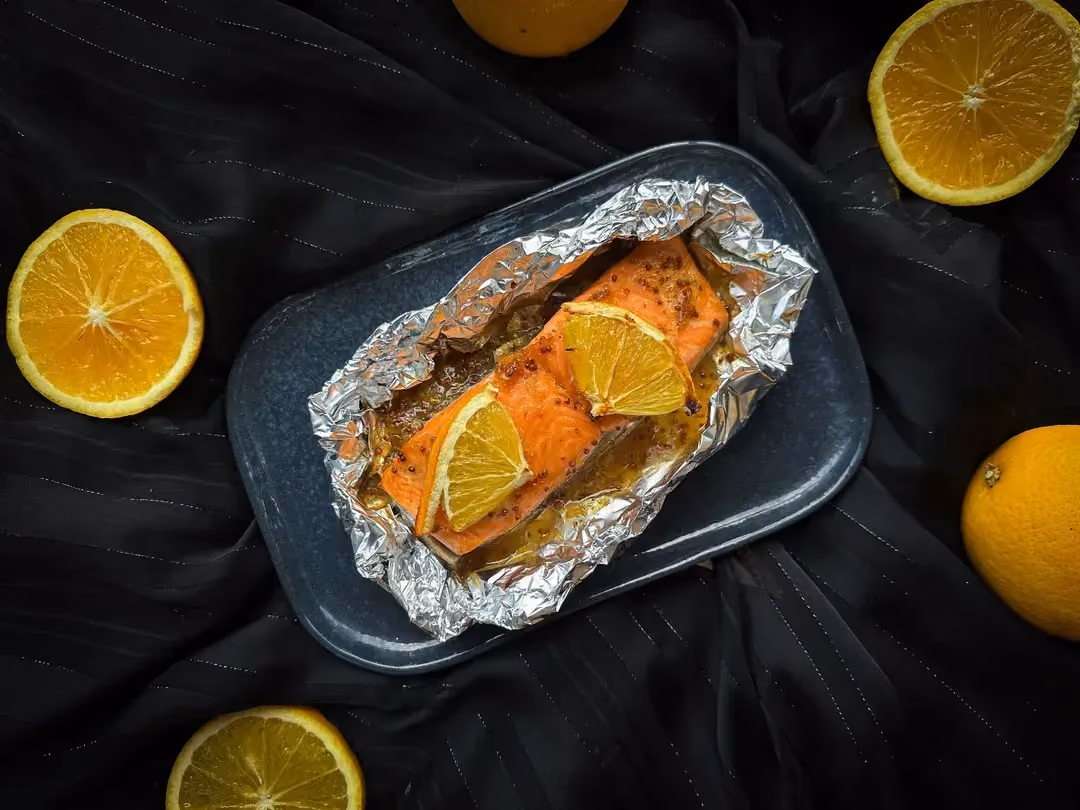 Sur une plaque de cuisson sont déposés trois pavés de saumon glacé à l'orange. Sur chaque morceau de saumon est déposé une tranche d'orange.