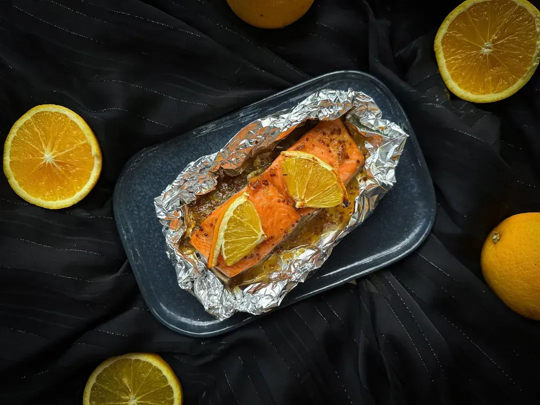 On a baking sheet, there are three orange-glazed salmon fillets. A slice of orange is placed on each piece of salmon.