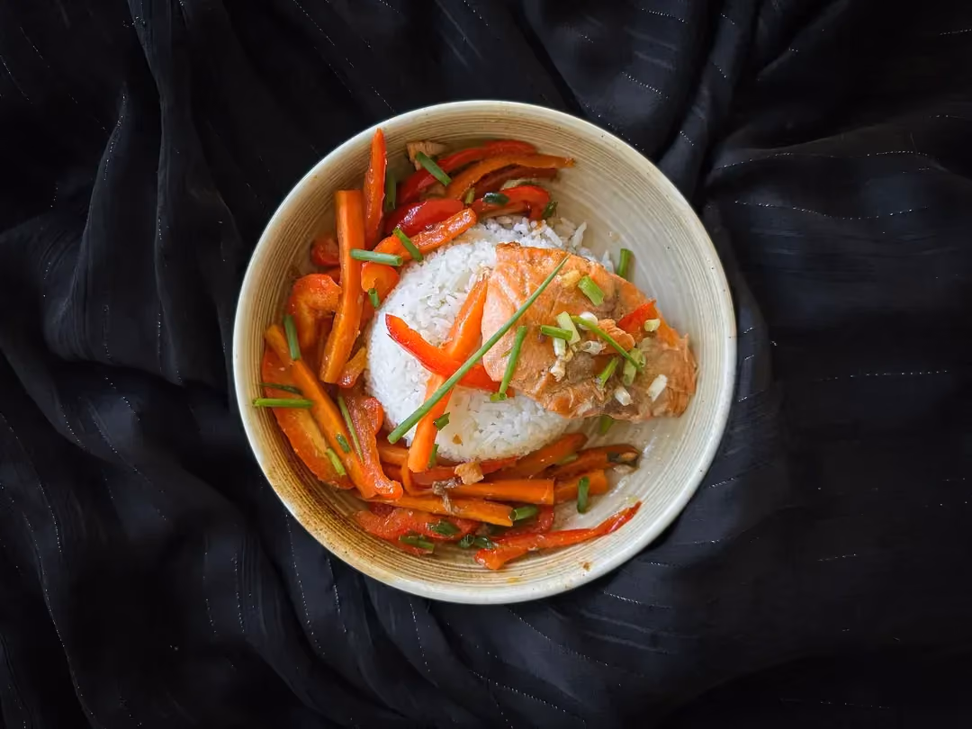 Dans un plat de couleur blanc se trouve un pavé de saumon caramélisé cuisiné façon teriyaki qui repose sur un lit à base de riz blanc.