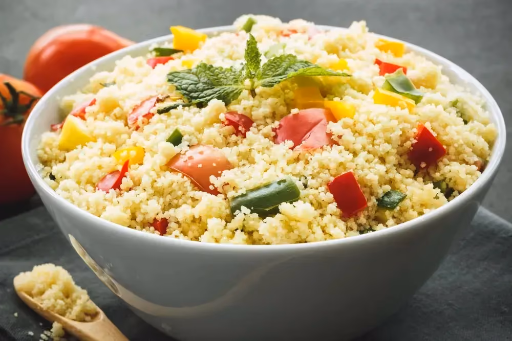 Dans un grand saladier de couleur blanc, se trouve la semoule accompagnée des légumes du soleil comme les tomates et les poivrons.
