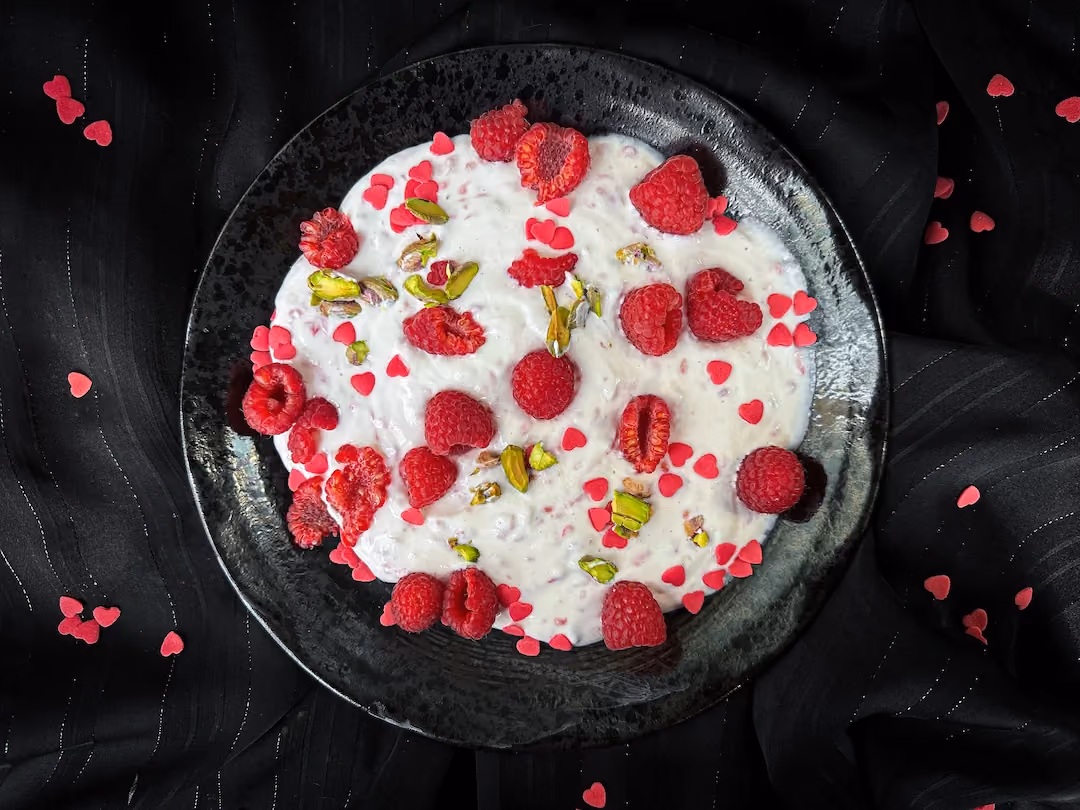 Posé sur un fond noir, un assiette noire contenant du Skyr aux framboises avec quelques pistaches concassées sur le dessus.
