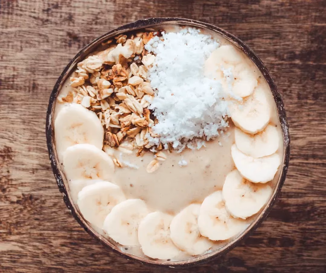 Dans un bol se trouve le smoothie. Sur le dessus se trouve une banane coupée en fines tranches, Des amandes concassées et du coco en poudre.