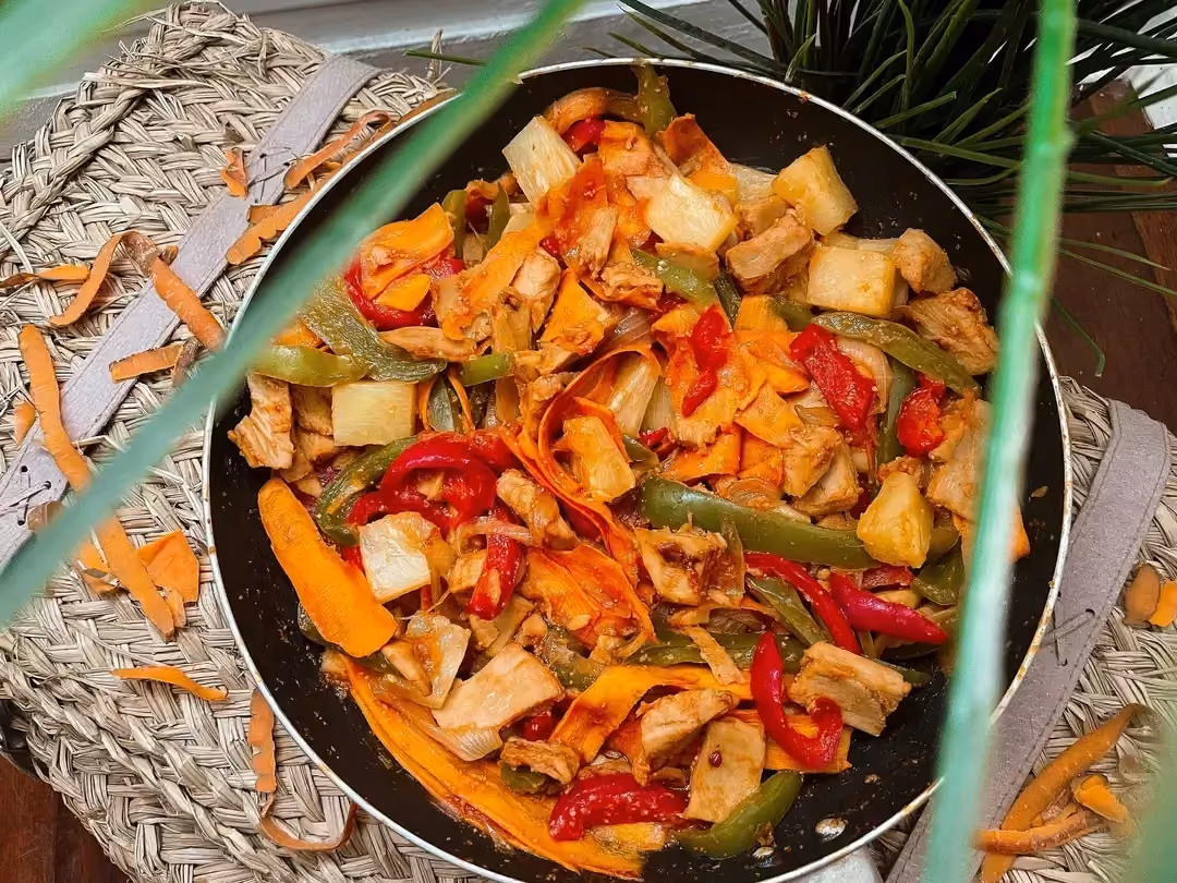 Le soja est cuisiné avec des poivrons, des carottes, de l'ananas et une sauce aigre-douce, le tout présenté dans une poêle au milieu de feuillage.
