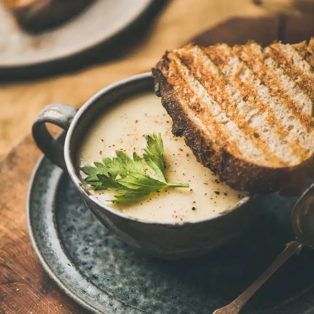 Chestnut and celeriac soup sits in a large mug. A thick slice of country bread partially covers the mug.