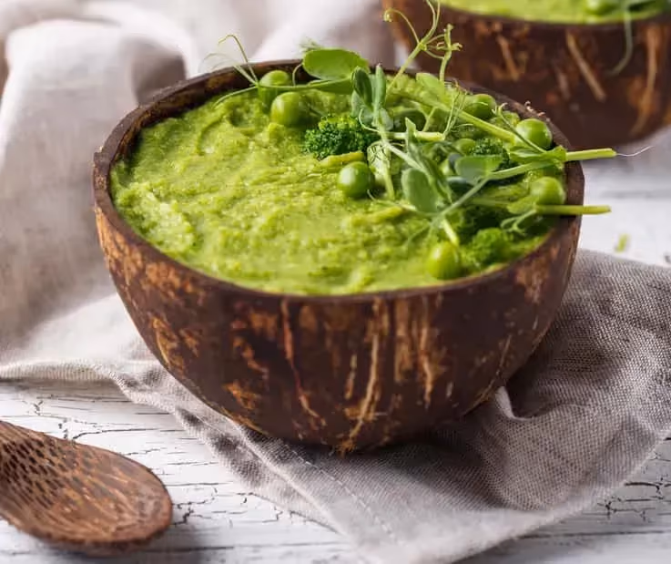 Broccoli soup is served in two fairly deep wooden bowls. A spoon made of the same material as the bowls is placed beside them.