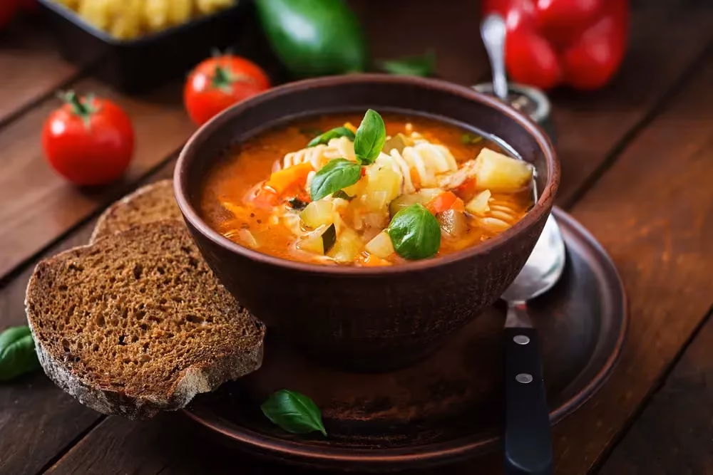 La soupe minestrone se trouve dans un bol de couleur marron. Une épaisse tranche de pain noir est à côté du bol. Des tomates sont posées sur la table.