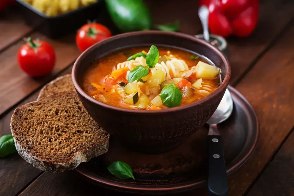The minestrone soup is in a brown bowl. A thick slice of dark bread is next to the bowl. Tomatoes are placed on the table.