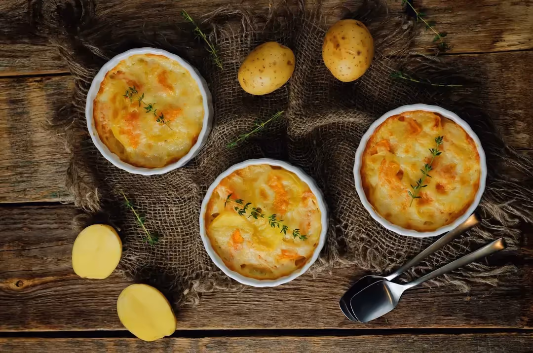 Présenté sur une table rustique, trois petits ramequins ronds contiennent ce Parmentier au saumon. Quelques pommes de terre sont disposées autour.