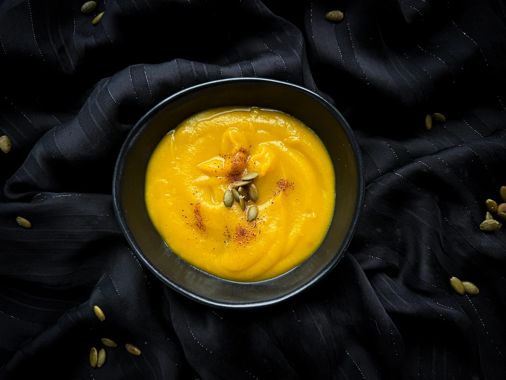 A small squash, hollowed out, serves as a bowl for the pumpkin, carrot, and orange soup. Two slices of bread and a small pumpkin are next to it.