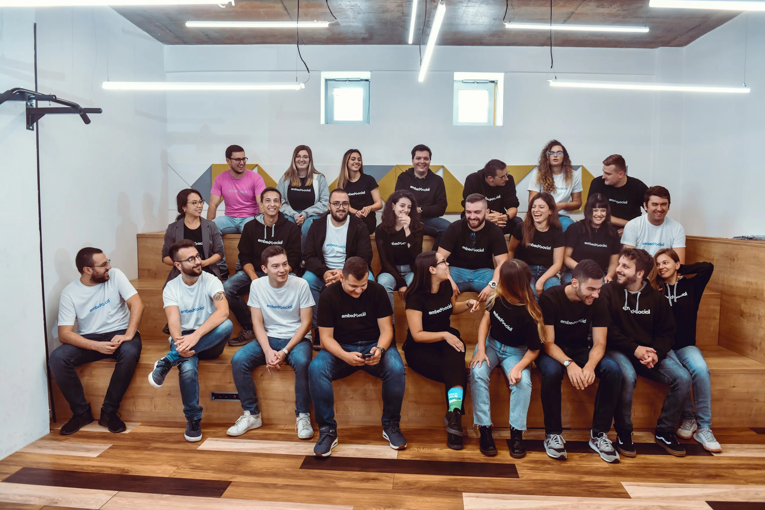 Group of 24 people casually dressed in branded embedSocial shirts sitting and smiling on wooden steps in a modern office space.