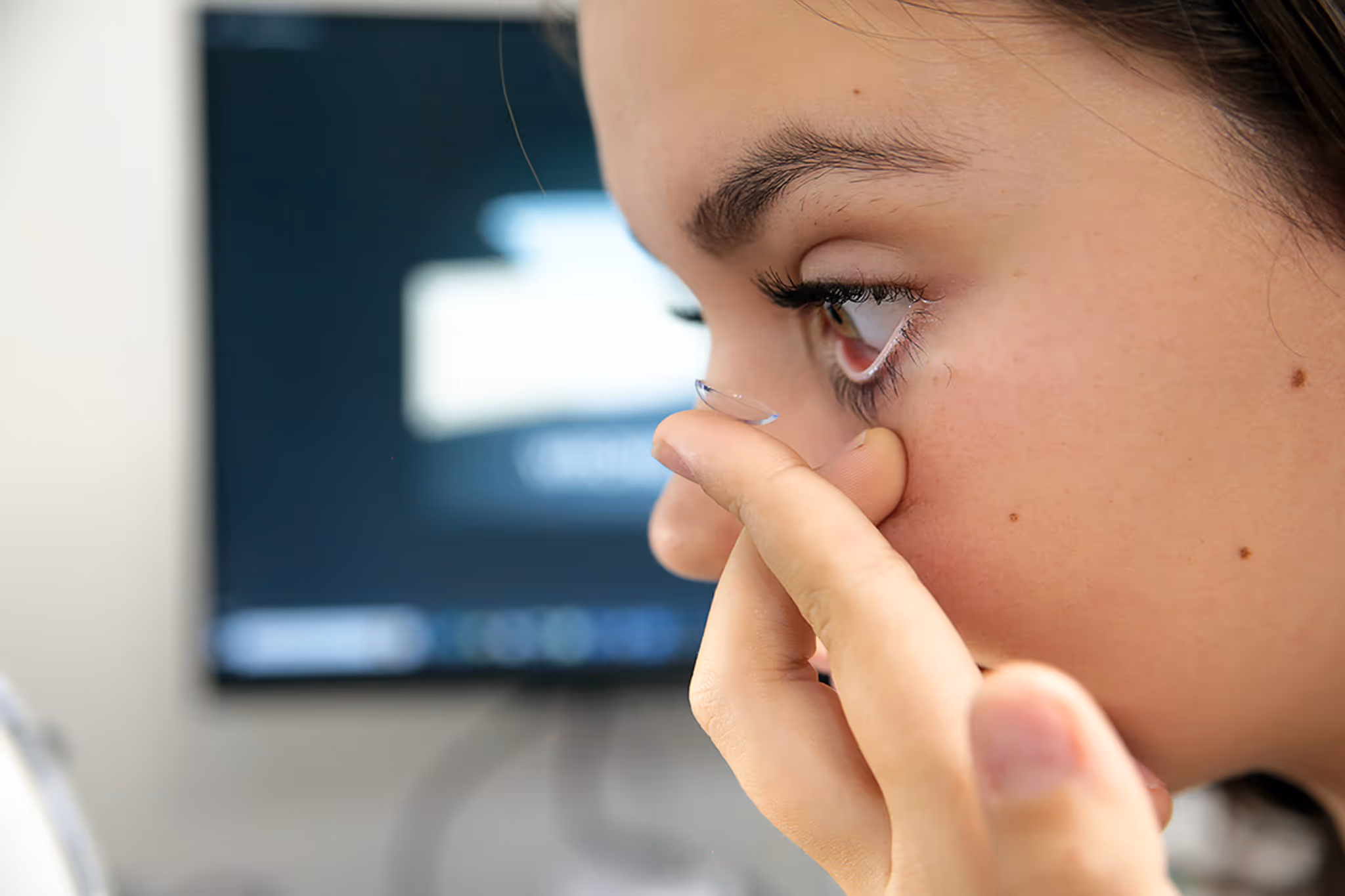 Close-up of person inserting contact lens with computer screen in background