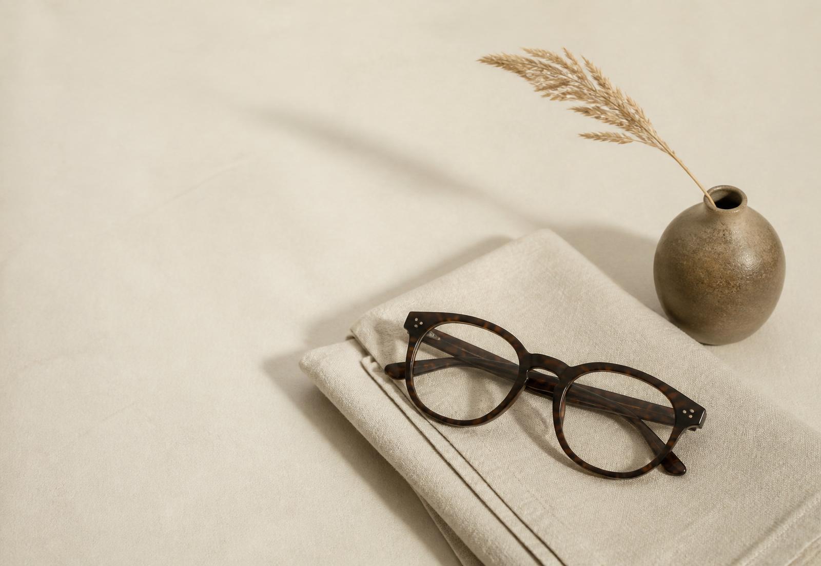 Brown tortoiseshell eyeglasses resting on a folded beige linen cloth next to a small round ceramic vase holding a single dried grass stem on a light beige background.