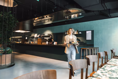 Eine Frau trägt ein Tablett mit Speisen in einem modernen Café mit dunklem Tresen und Sitzbereich.