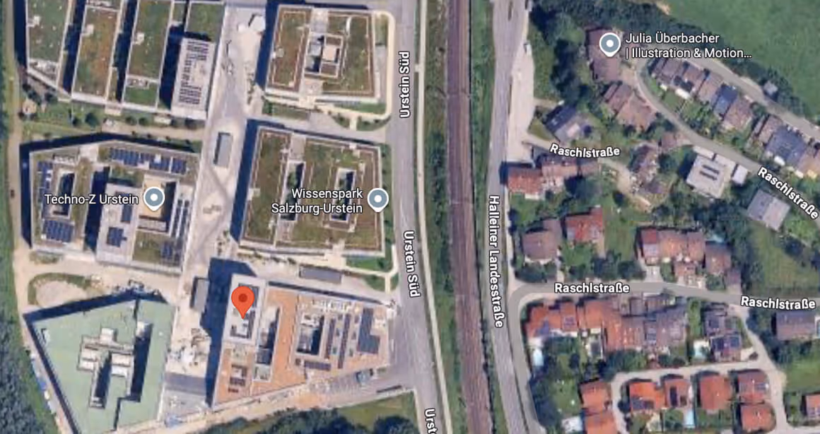 Satellitenaufnahme mit Technologie- und Wissenschaftsparkgebäuden auf der linken Seite und Wohnhäusern rechts, getrennt durch eine Bahnstrecke in Urstein, Salzburg.