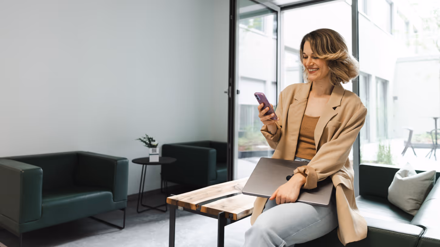 Frau mit beige Jacke, die lächelnd auf ihr Smartphone schaut und einen Laptop auf dem Schoß hält in einem modernen Büro mit Sitzbereich.