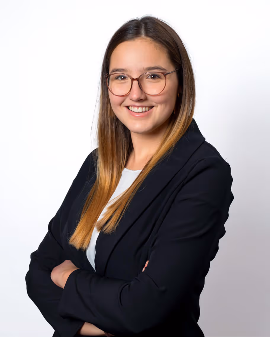 Junge Frau mit Brille und langen braunen Haaren trägt schwarzen Blazer und weißes Shirt, steht vor weißem Hintergrund mit verschränkten Armen und lächelt.