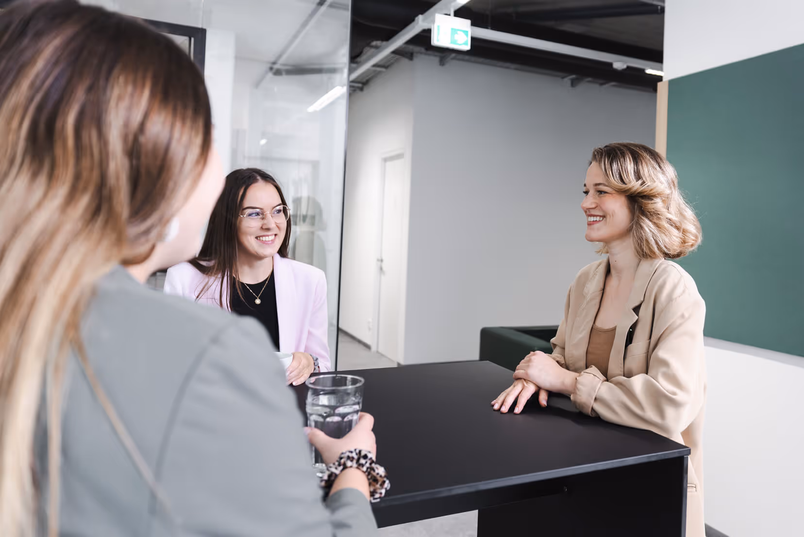 Drei Frauen unterhalten sich lächelnd bei einem hohen Tisch in einem modernen Büro mit Glaswänden.
