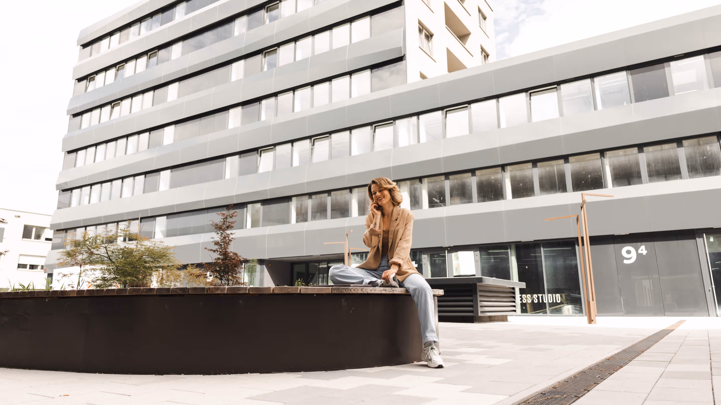 Frau in Beige-Jacke sitzt im Schneidersitz auf einer Bank vor einem modernen Bürogebäude und telefoniert lächelnd.