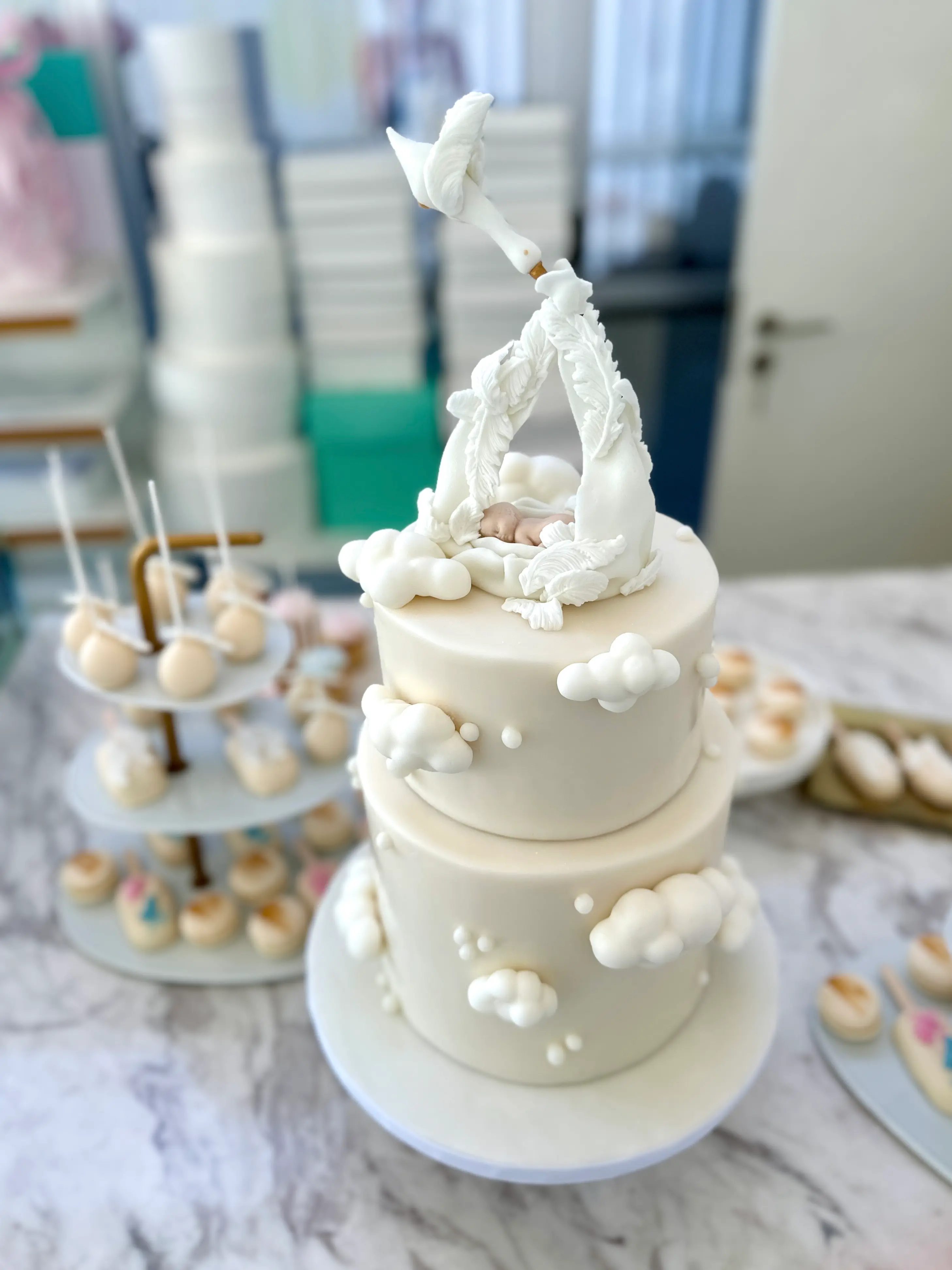 Elegant gender reveal cake with all-white design, two-tier fondant cake decorated with soft clouds and a sculptural baby topper, luxury dessert table setup in cyprus
