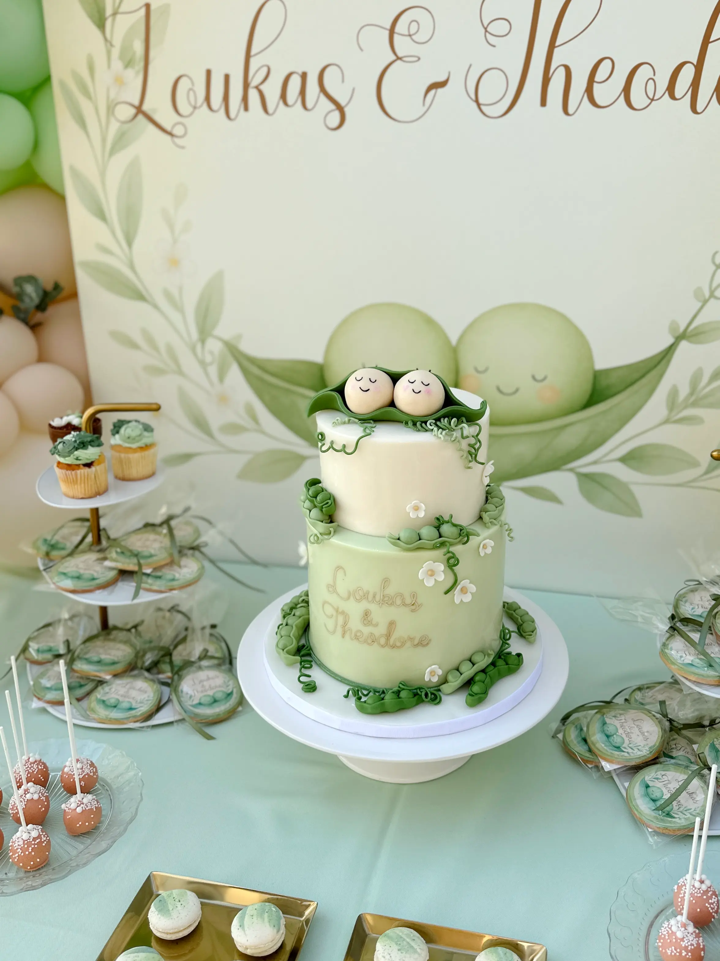 Elegant christening cake and dessert table for twin boys with “two peas in a pod” theme, two-tier pastel green cake with baby toppers in a pea pod, custom cookies, cupcakes, and coordinated sweets display in soft green tones