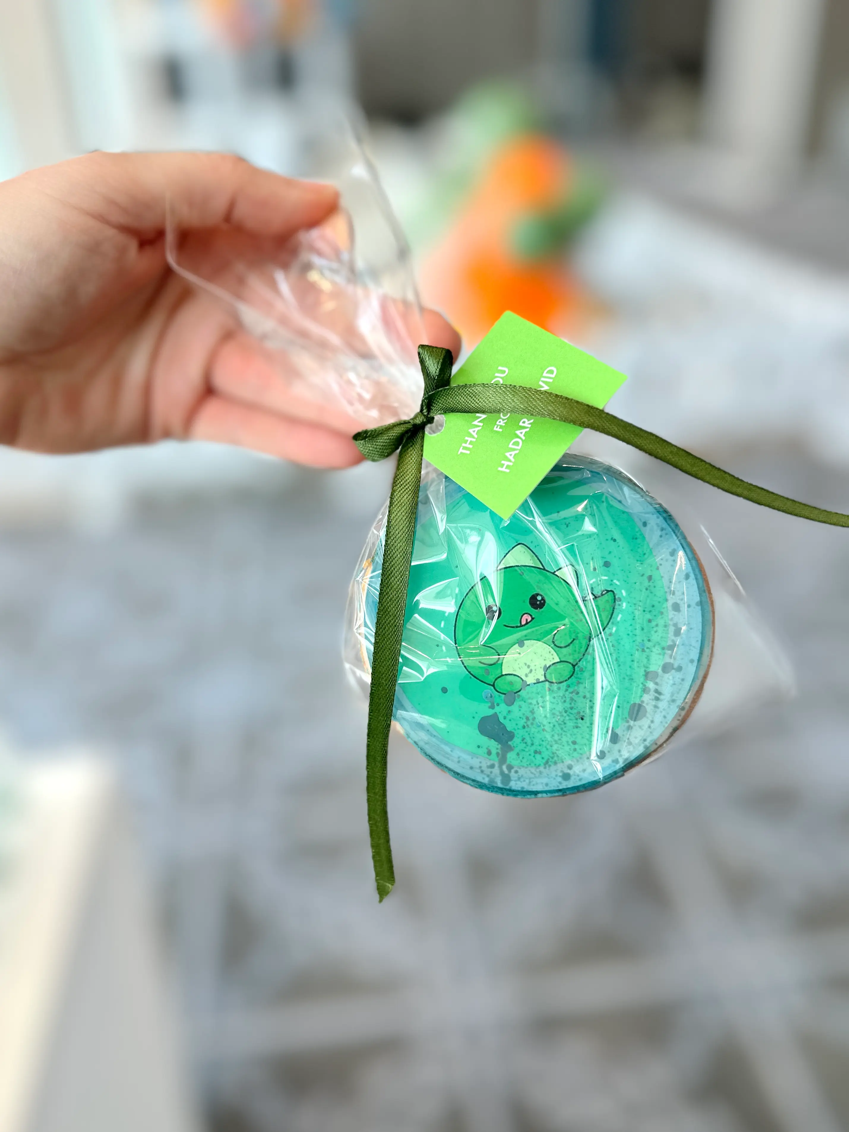 Individually wrapped dinosaur-themed cookie favour held in hand, decorated in green tones with a cute baby dinosaur design, tied with a green ribbon and personalised thank-you tag for a first birthday celebration