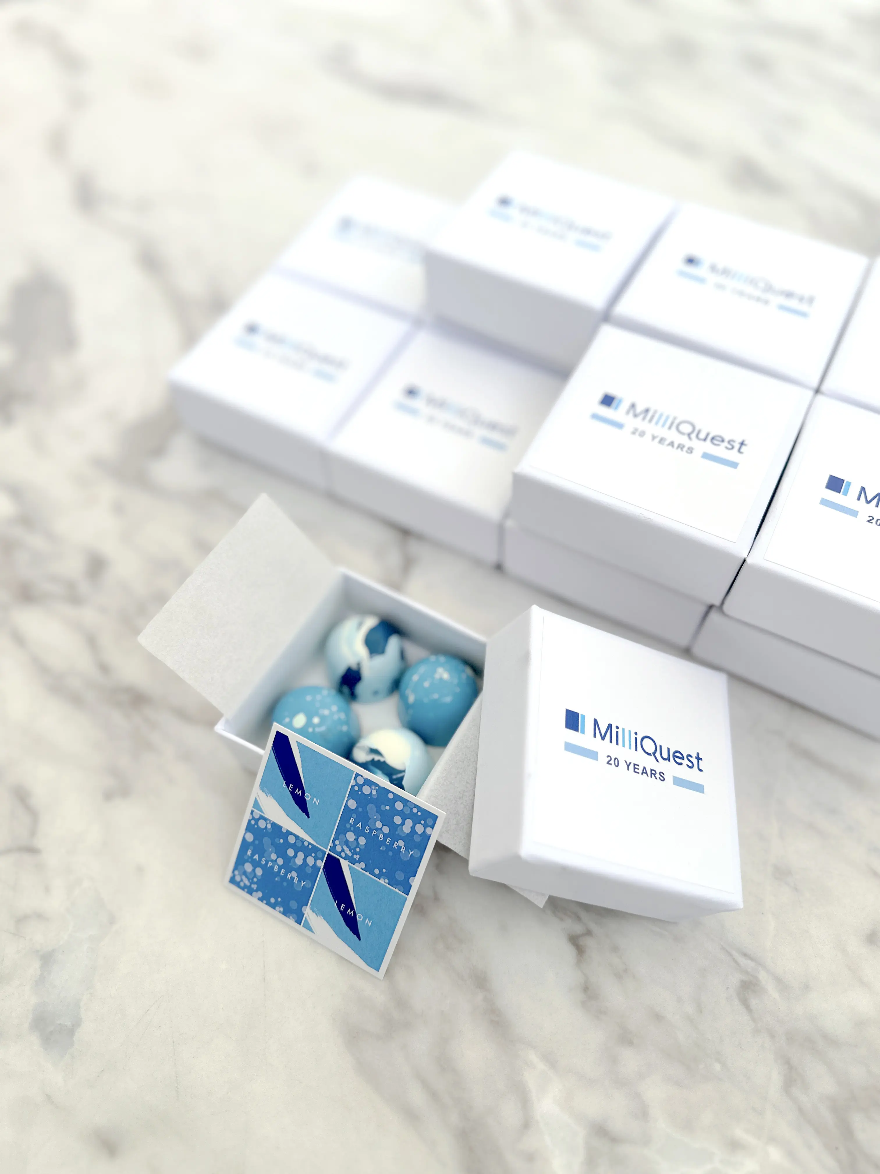 Elegant corporate chocolate gifts in minimalist white boxes with custom logo, containing artisanal blue-toned bonbons, styled for a company milestone event and premium client gifting
