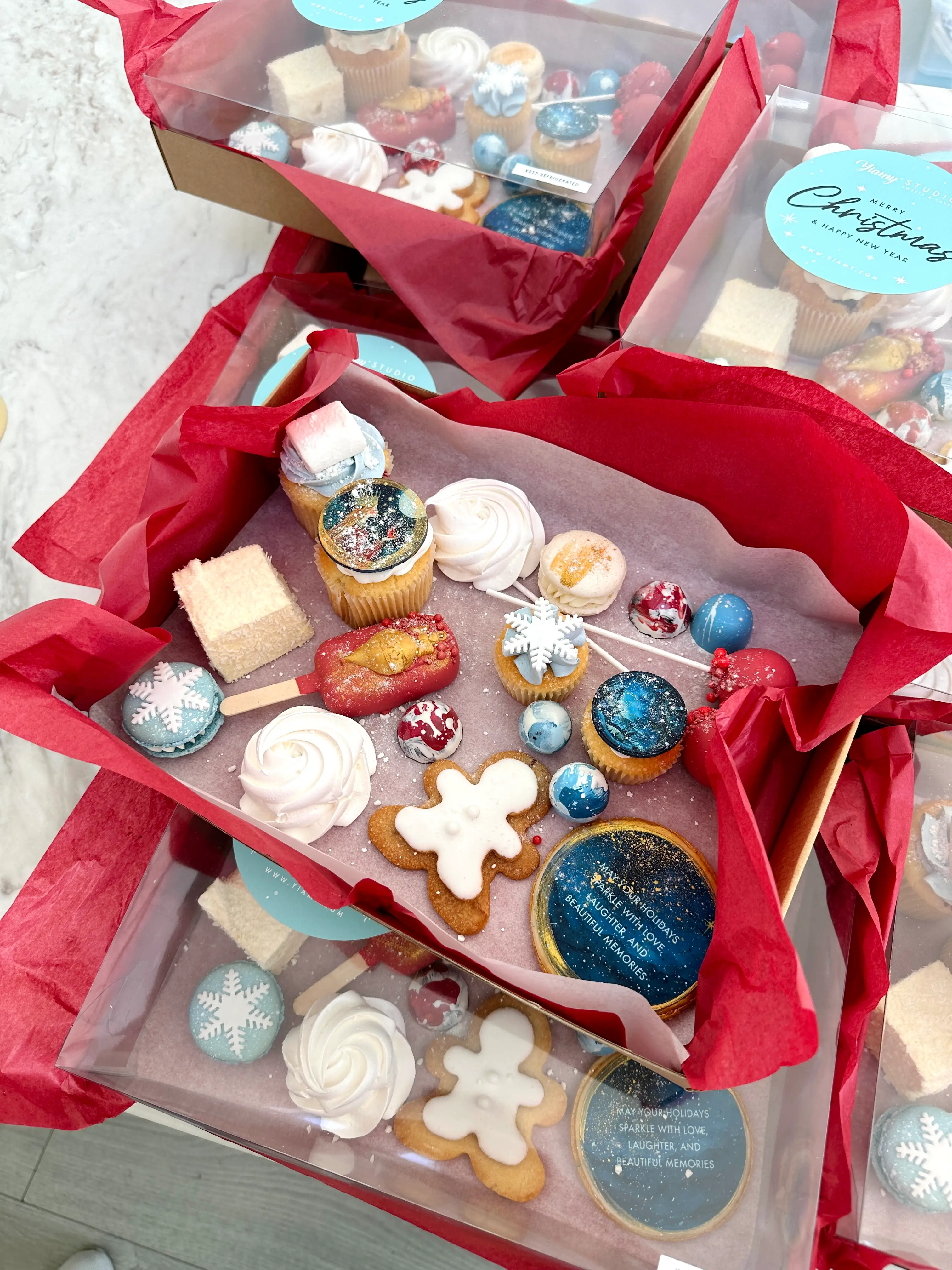 Luxury corporate christmas dessert platter gift box filled with festive treats including decorated cookies, cupcakes, macarons, meringues, and chocolate bonbons in red, white, and blue tones, elegantly packaged with tissue paper for holiday gifting Cyprus