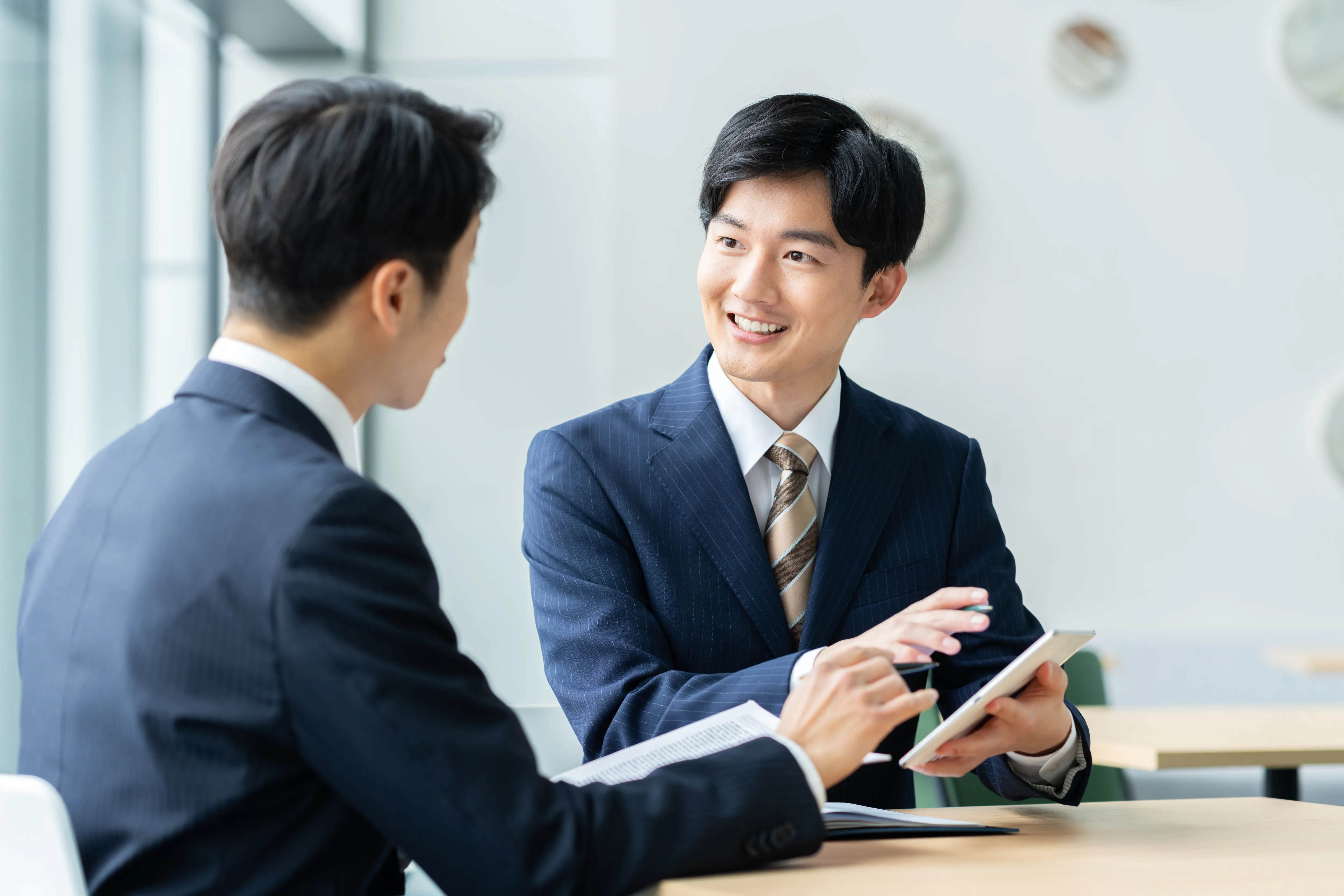 スーツを着た二人の男性が明るいオフィスでタブレットと書類を使いながら会話している。