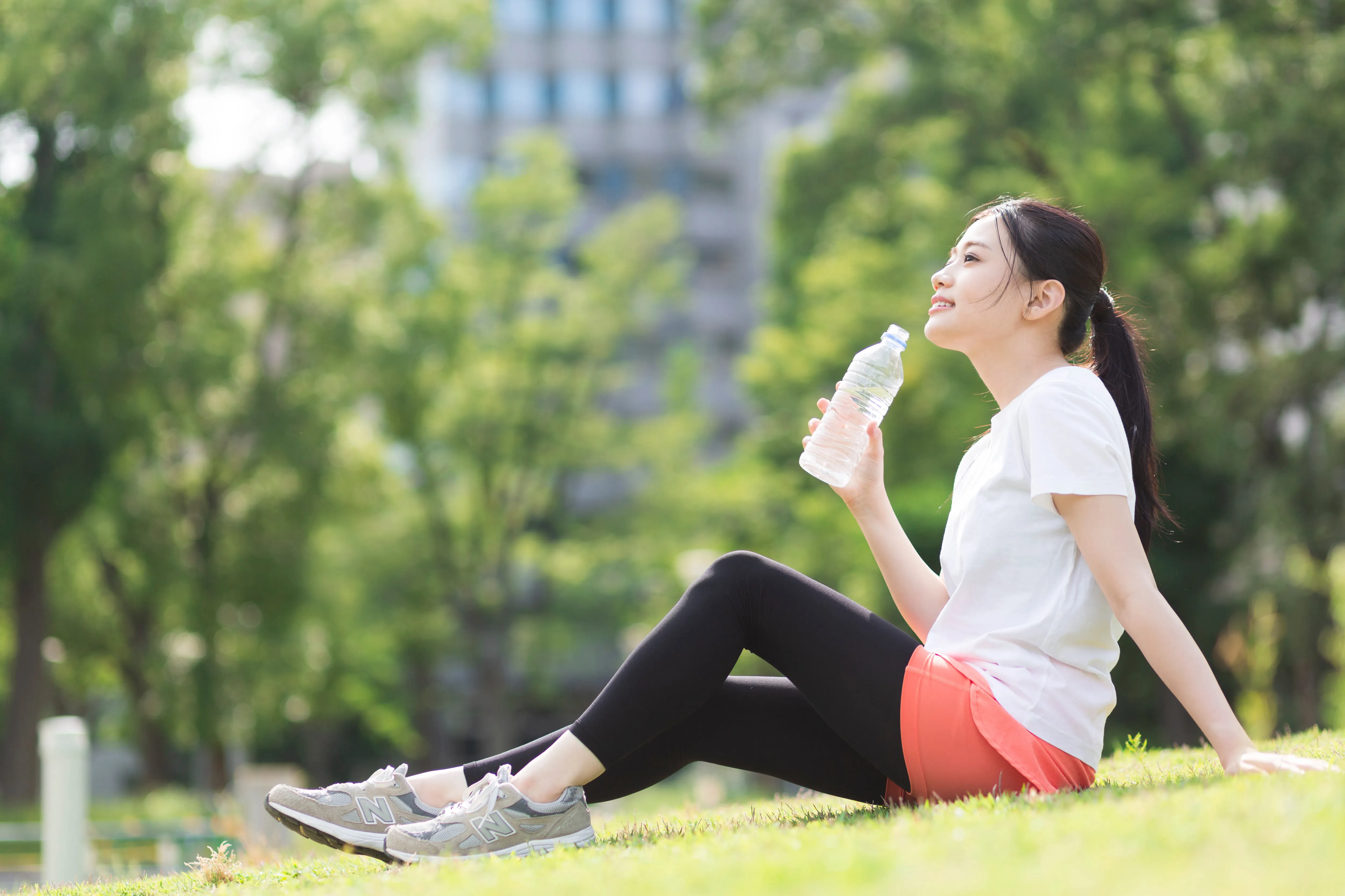 公園の芝生に座り、ペットボトルの水を飲もうとしている女性。