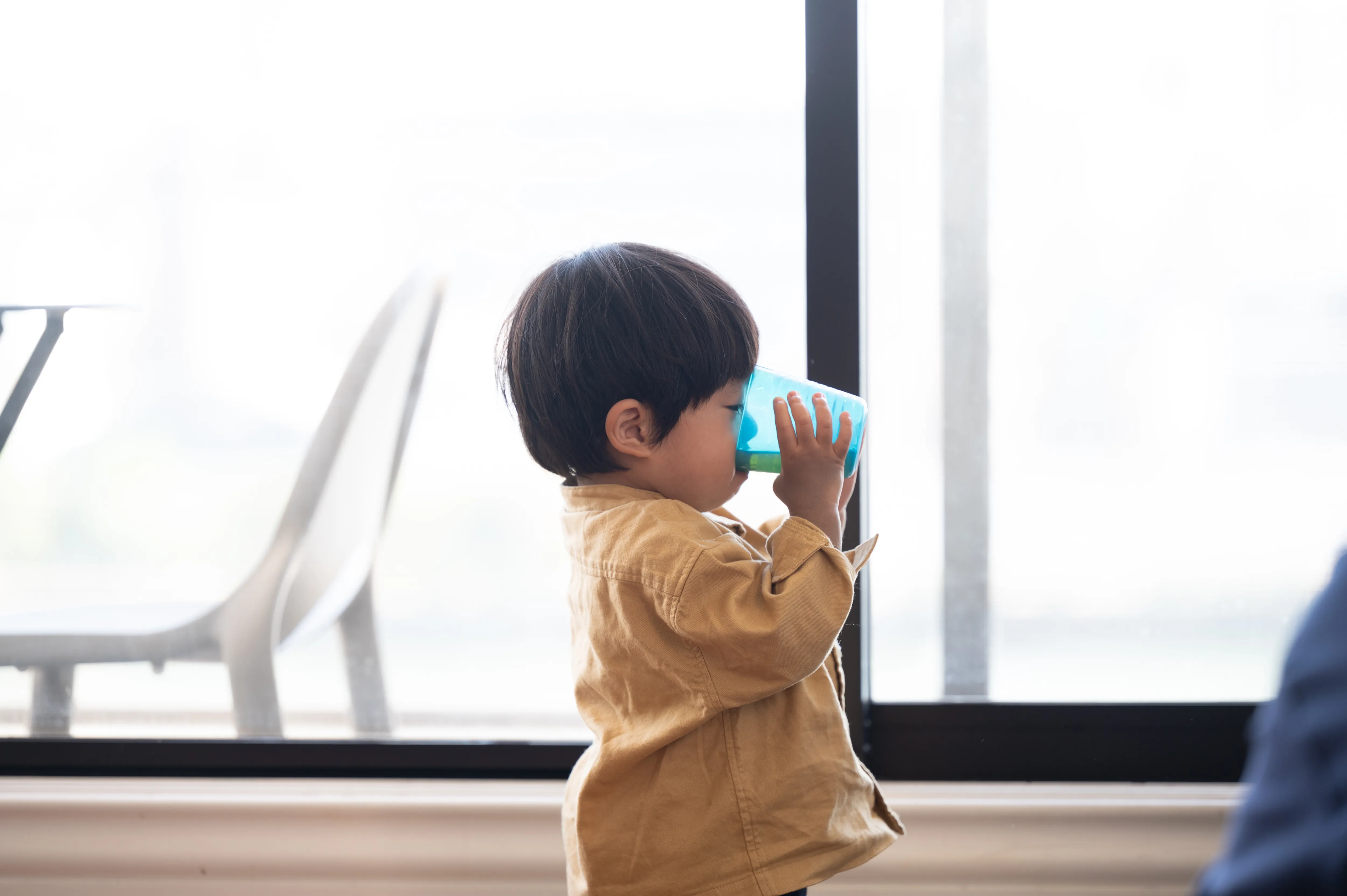 青いコップで飲み物を飲んでいるベージュのジャケットを着た幼い男の子。