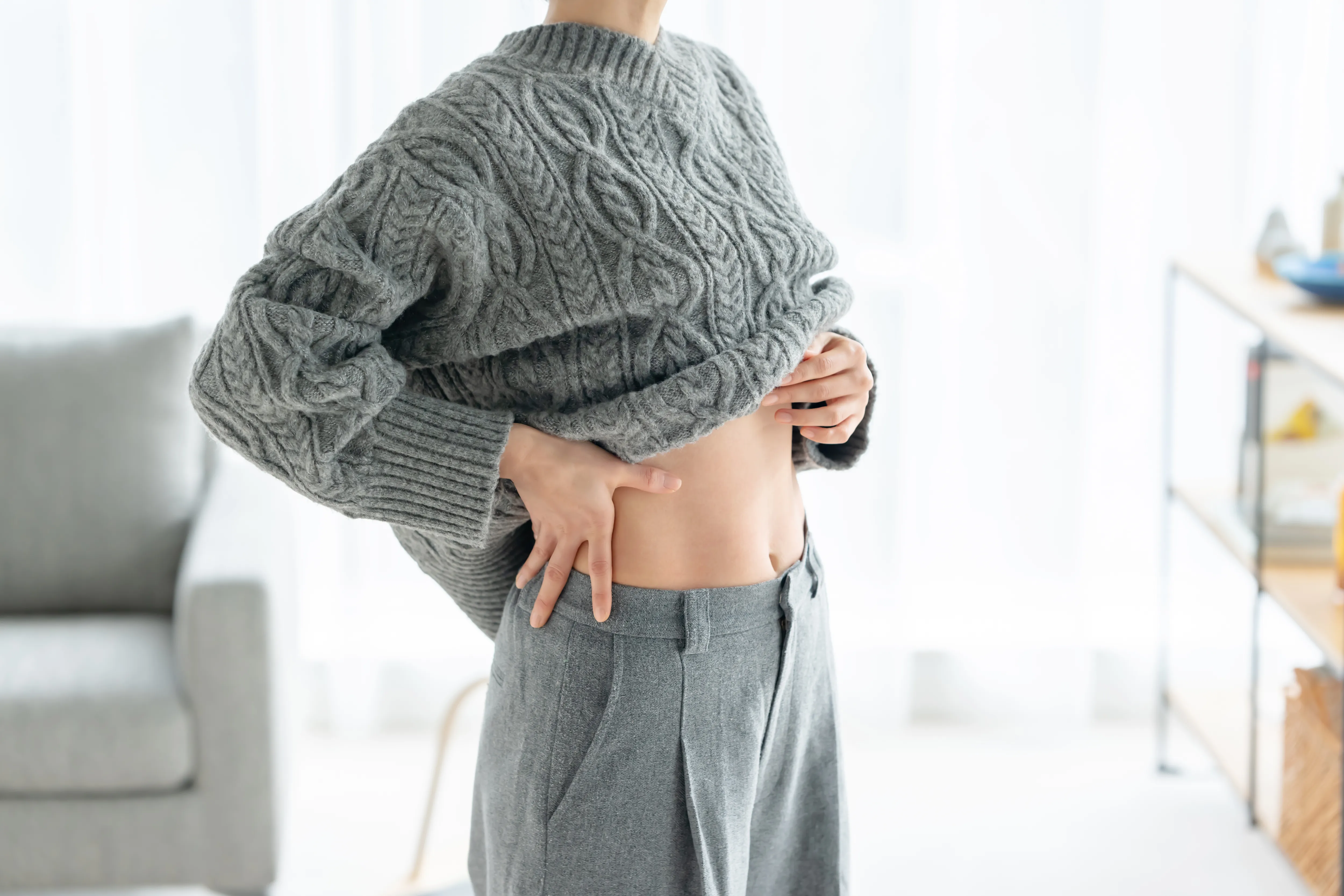 グレーのケーブルニットセーターをめくり、お腹の側面を触る女性。