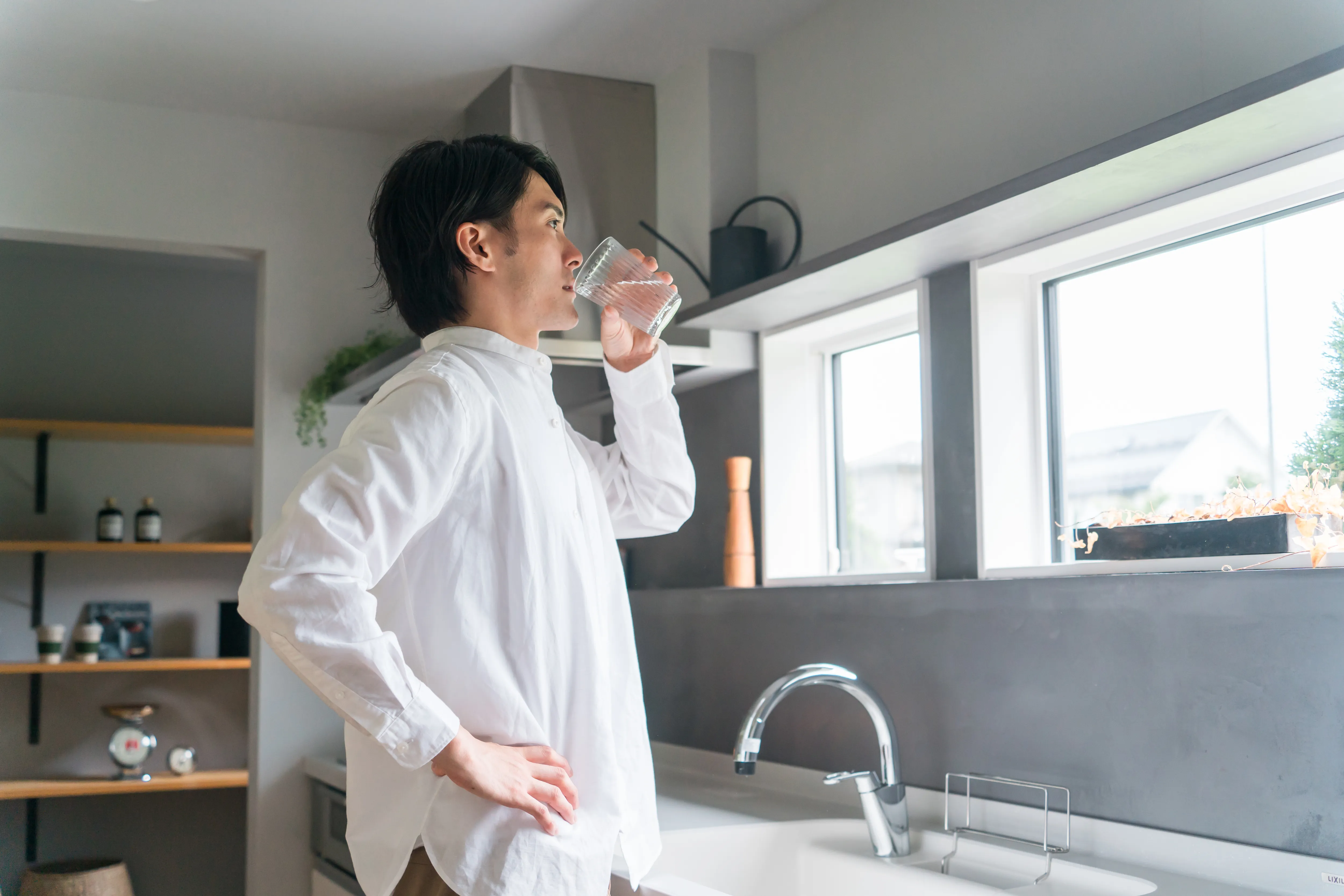キッチンで窓の外を見ながらグラスの水を飲む白いシャツの男性。