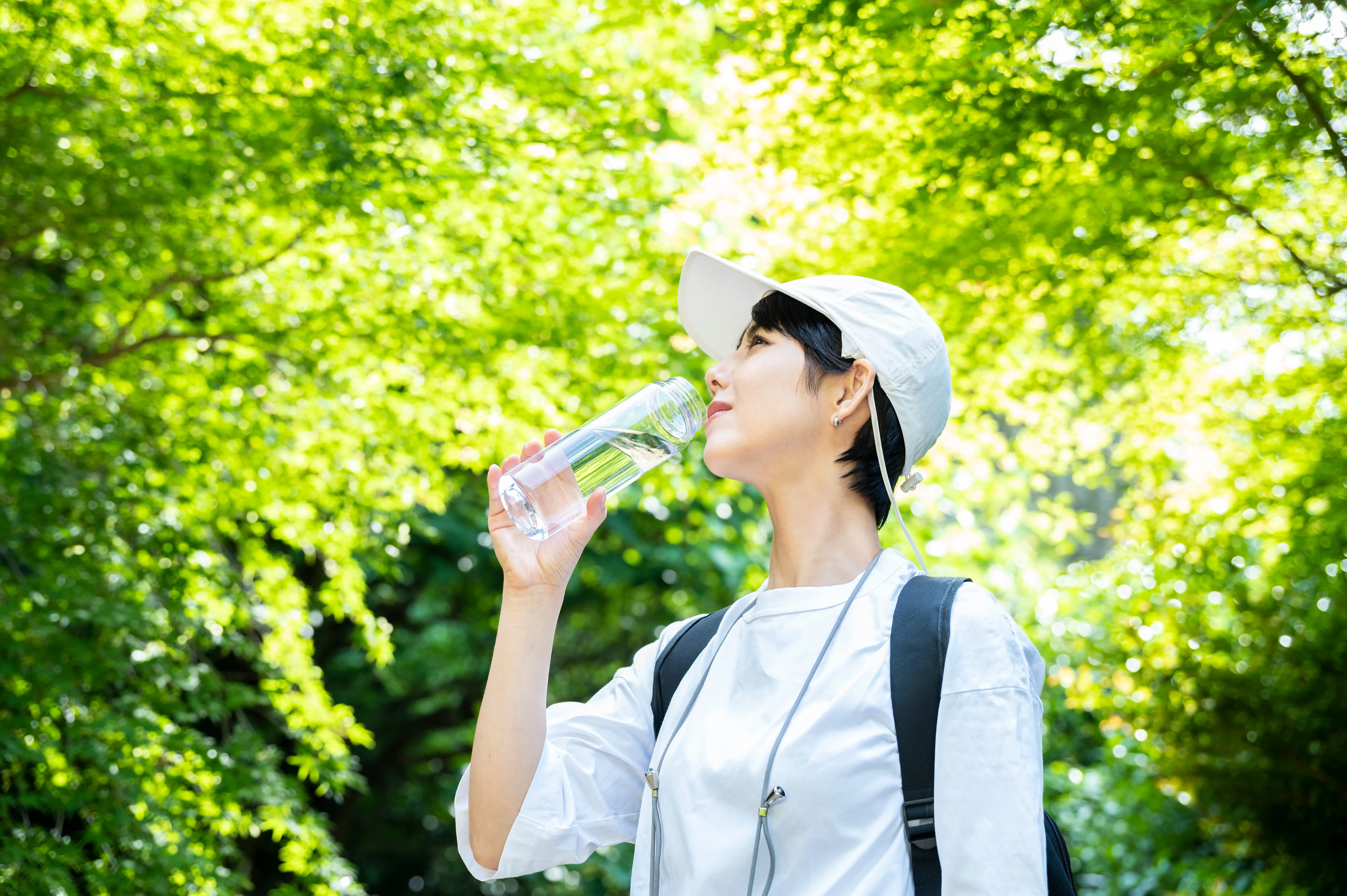 白い帽子をかぶり白い服を着た女性が緑の木々の中で水の入ったボトルを飲んでいる。