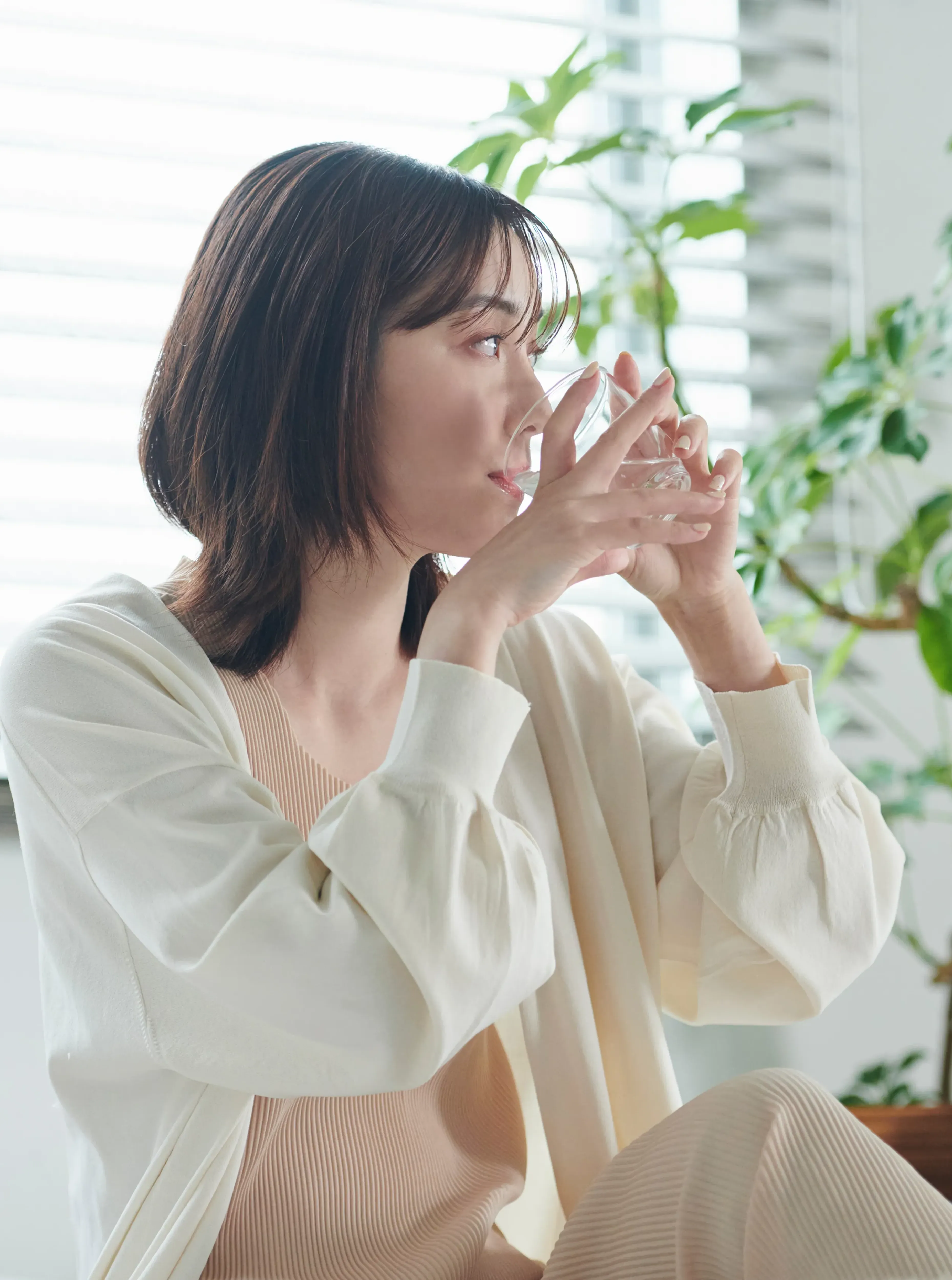 窓辺でグラスの水を飲んでいるベージュの服を着た女性。