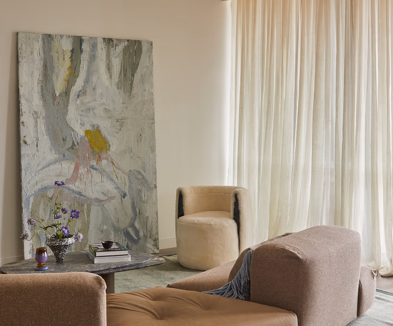 Modern living room with a textured abstract painting leaning against the wall, a marble coffee table with books and flowers, a beige fluffy chair, and a brown sofa near sheer curtains.