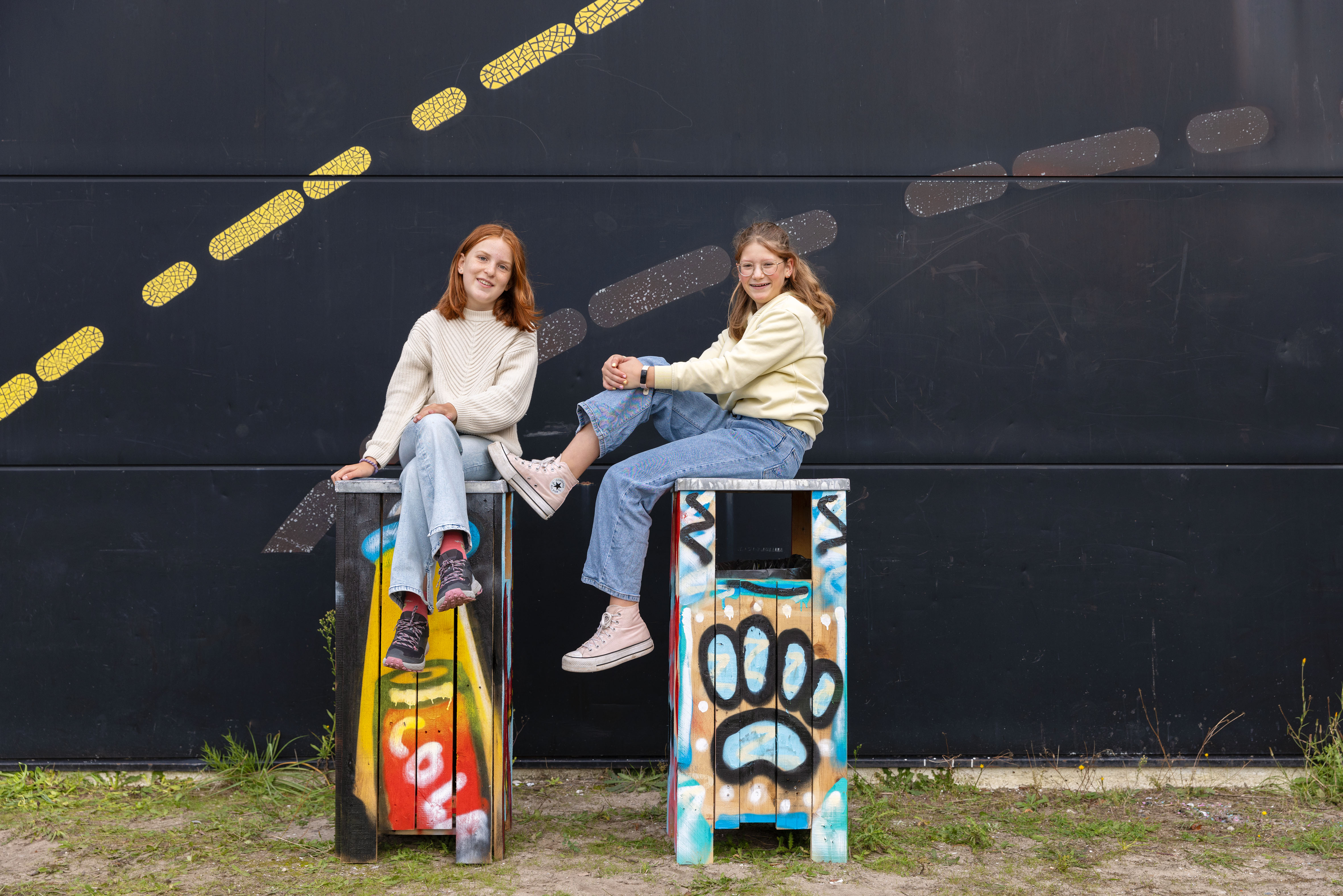 Two girls sitting on graffiti-covered wooden stools in front of a black wall with yellow and gray dashed lines.