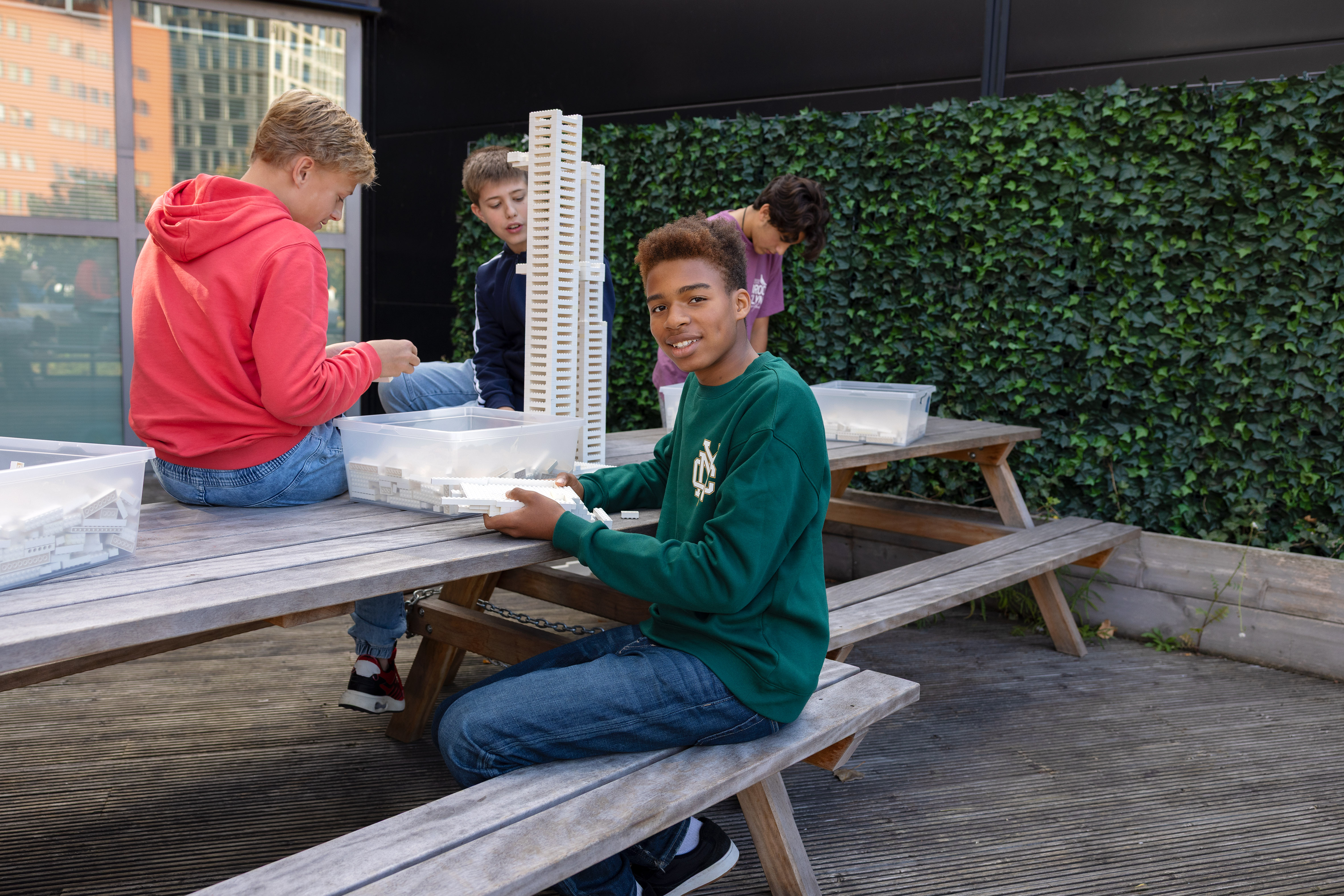 Four boys sitting at wooden picnic tables outdoors, building a tall structure with white plastic construction blocks.