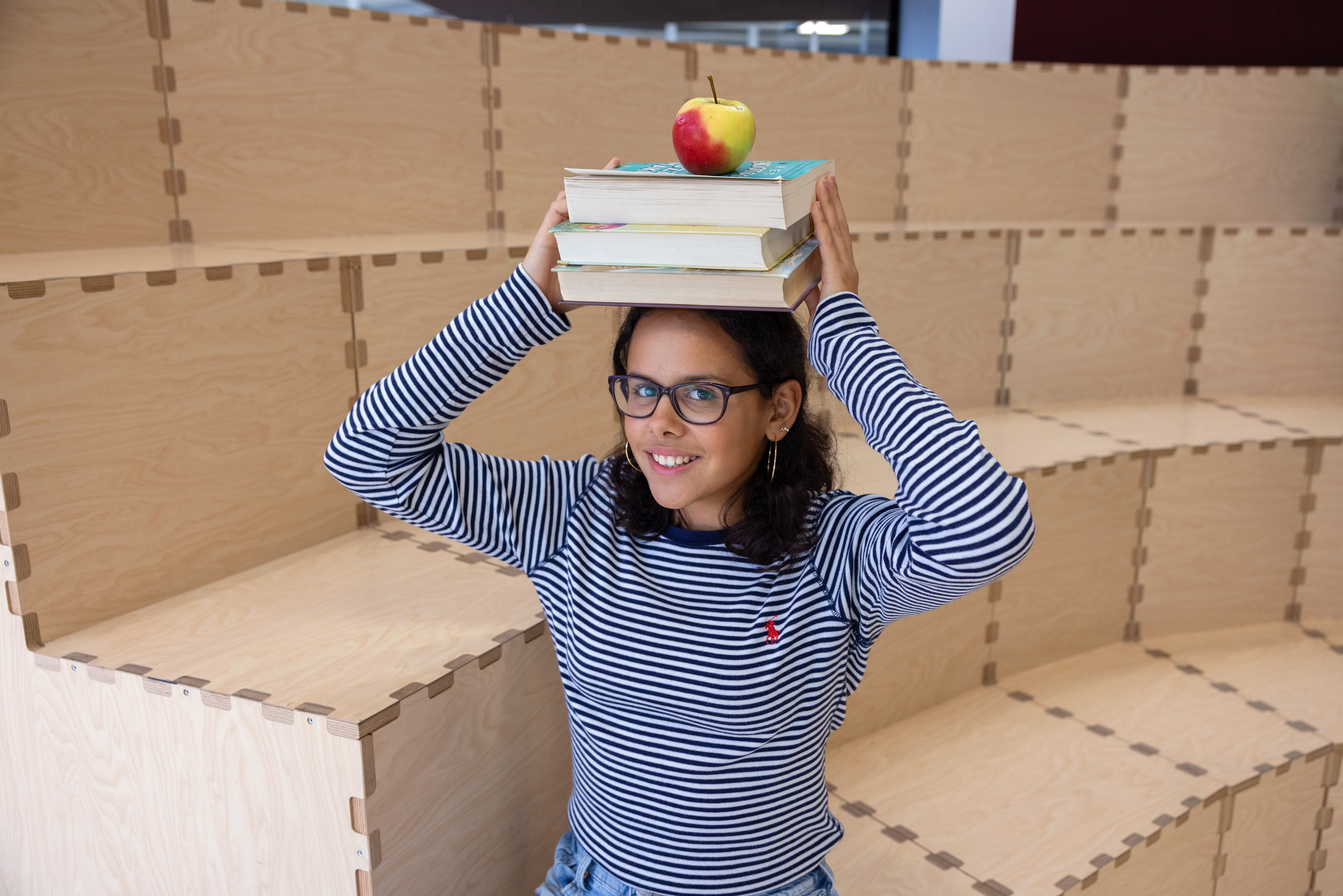 Smiling girl with glasses balancing a stack of books and an apple on her head while sitting on wooden tiered seating.