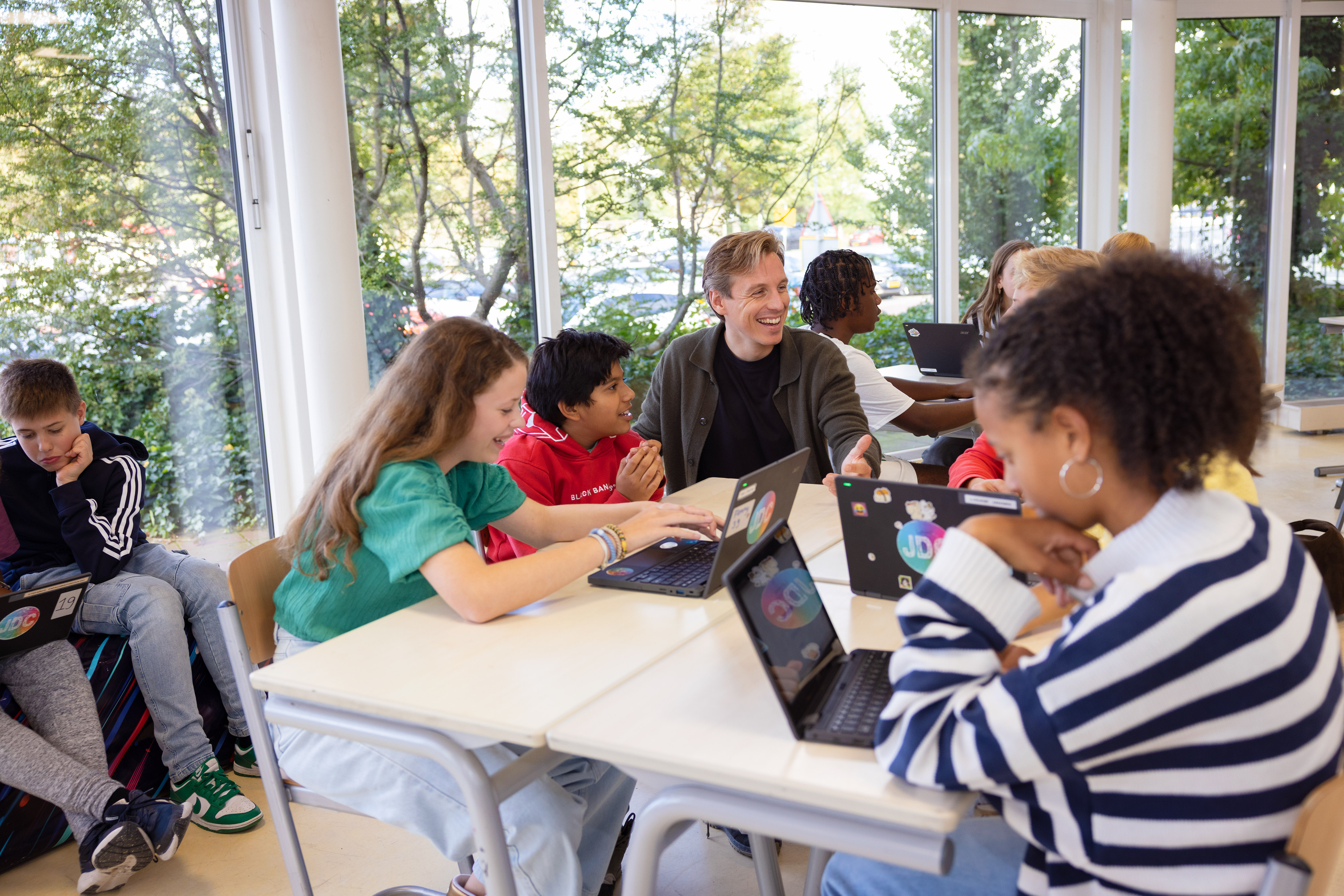 Group of children and a teacher working and interacting around laptops at a classroom table with large windows and greenery outside.
