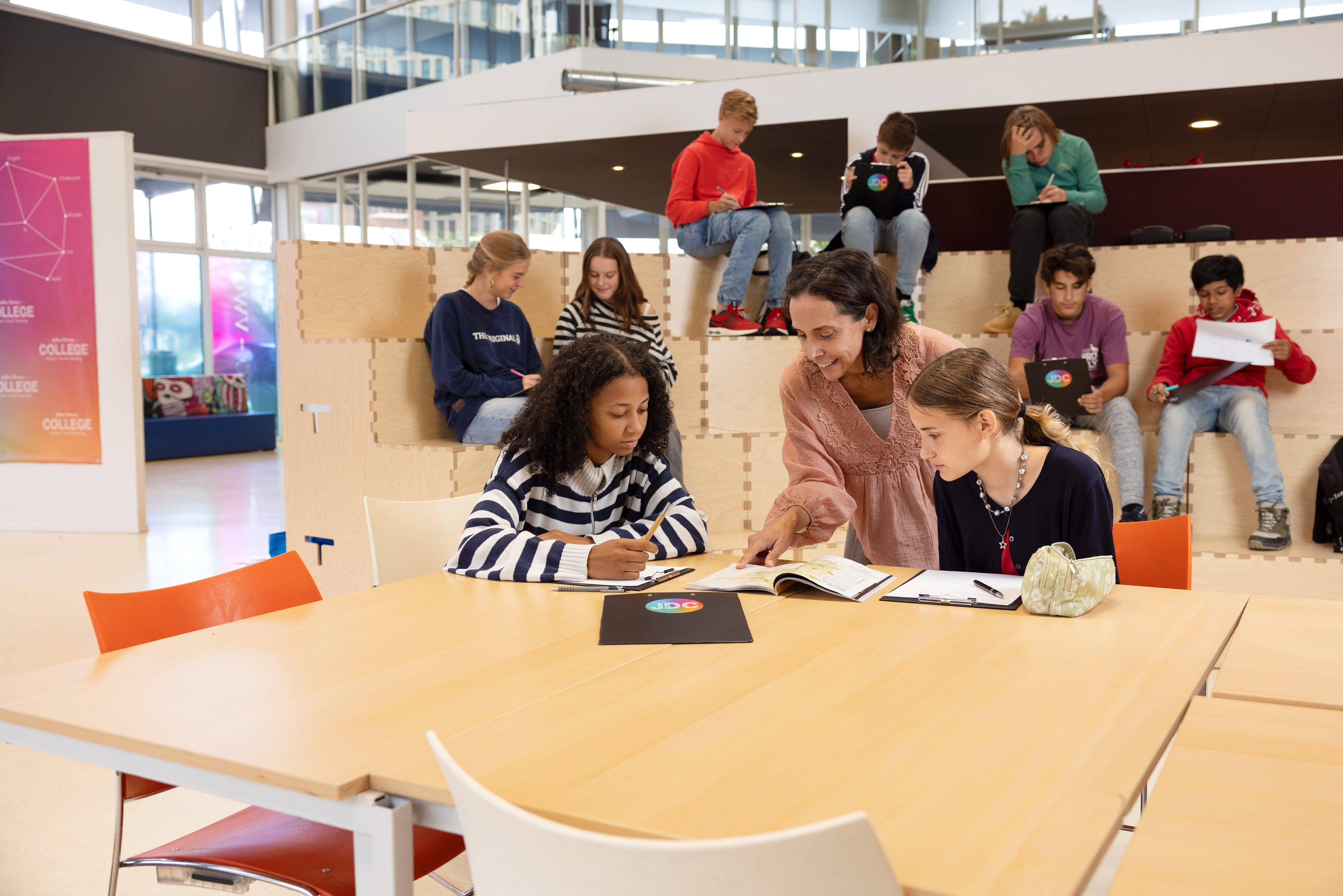 Teacher helping two students with a book at a table while other students study in the background.