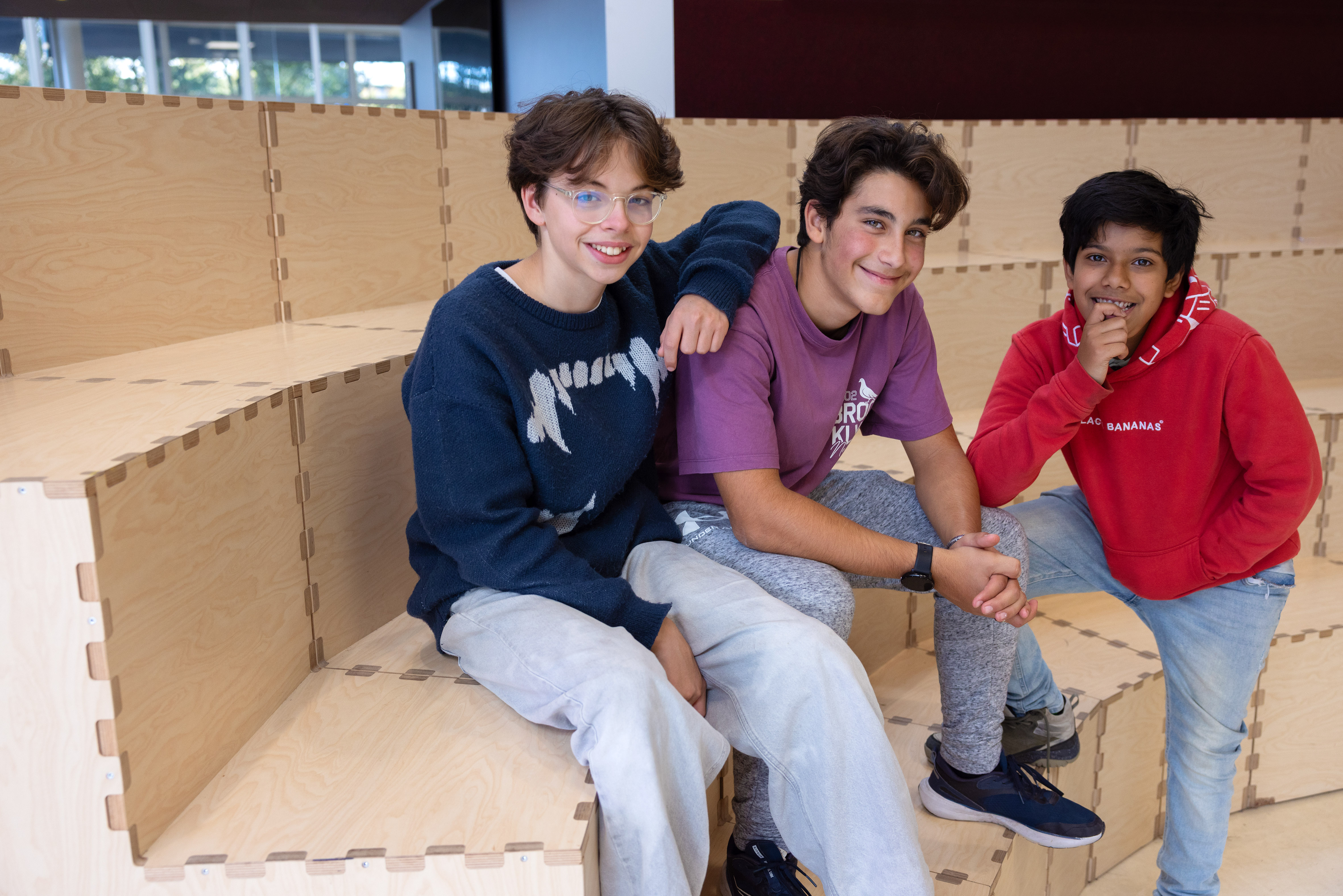 Three smiling boys sitting and posing casually on wooden tiered seating indoors.