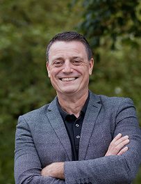 Smiling middle-aged man with short dark hair wearing a gray blazer and black shirt, standing outdoors with arms crossed.